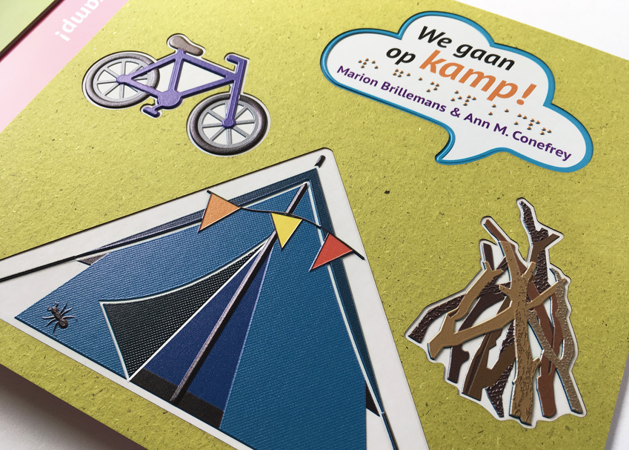 Voorkant 'We gaan op kamp’ opgebouwd uit een toplaag van geurend graspapier met daaronder de titel in braille en een tactiele illustratie.